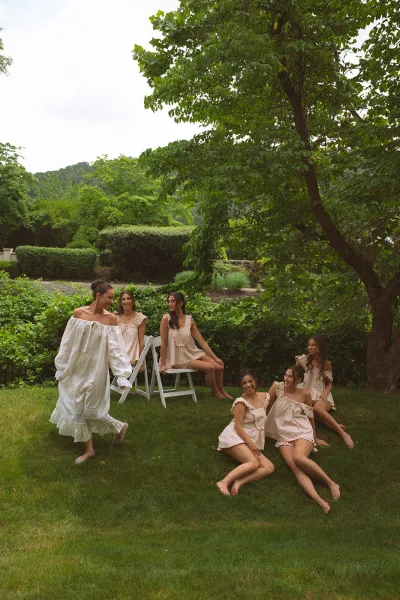 Bridal party portrait of a bride with bridesmaids, the bride in a white off-shoulder dress, gathered on a garden lawn by trees.
