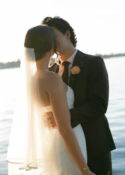 Couple portrait of a wedding kiss moment by tranquil water and distant trees, the bride's long bridal veil trailing as the groom holds her close.