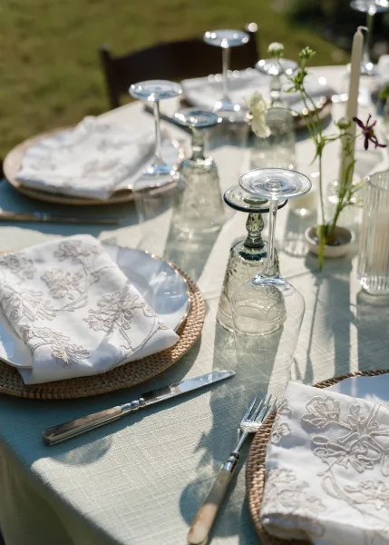 Reception tablescape on an outdoor reception table with embroidered napkins, set on a grass lawn with a wooden chair visible in the background.