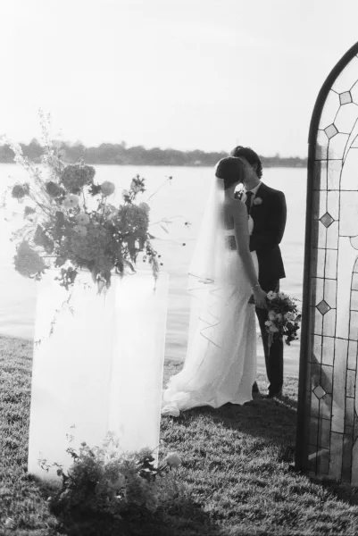 Wedding kiss portrait of a bride and groom kiss beneath a stained glass backdrop at a lakeside ceremony, framed by soft shoreline and sky.
