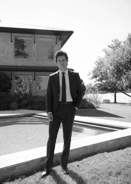 Groom portrait in black and white, standing in a suit beside a house exterior with windows and a pool on a lawn with trees.
