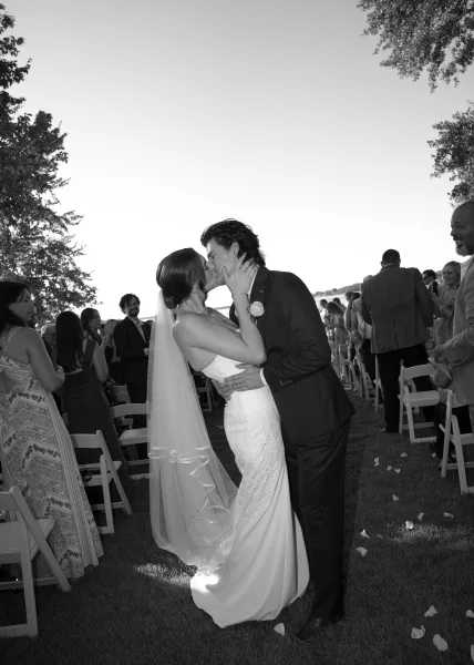 Wedding kiss portrait capturing a recessional kiss as newlyweds embrace on a flower petal–lined ceremony aisle between rows of folding chairs.