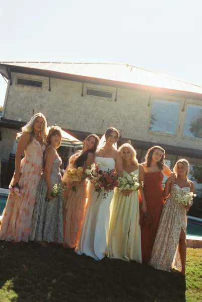 Bridal party portrait in mix and match bridesmaid dresses, holding bouquets beside a stone house and swimming pool on a lawn.