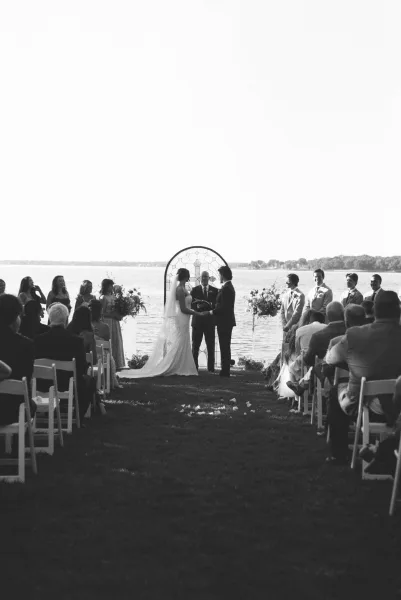 Ceremony moment at an outdoor wedding ceremony as the bride and groom face each other beneath a round arch on a lakeside lawn under open sky.