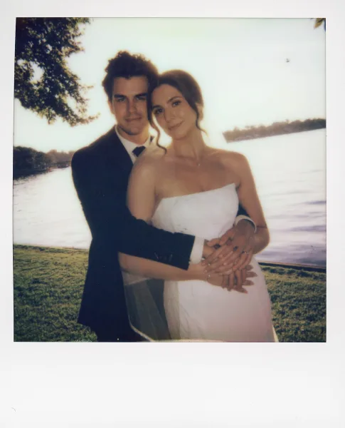 Couple portrait of a bride and groom embrace by a tranquil lake, her veil catching light as they stand on a grassy lawn framed by trees.