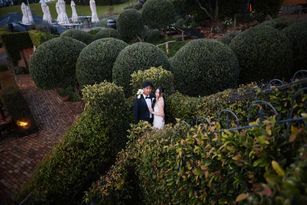 Couple portrait in a garden wedding portrait setting, the bride holds a white lily bouquet as they embrace amid manicured topiary hedges.