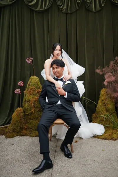Couple portrait with bride standing behind groom, her long tulle veil draping as she rests her hands on his shoulders against a velvet curtain.