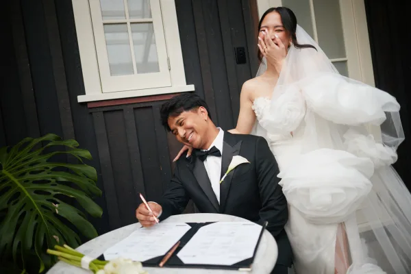 Marriage license signing as the groom signs the certificate beside his bride at a marble table, with a dark wood wall and monstera leaves behind.