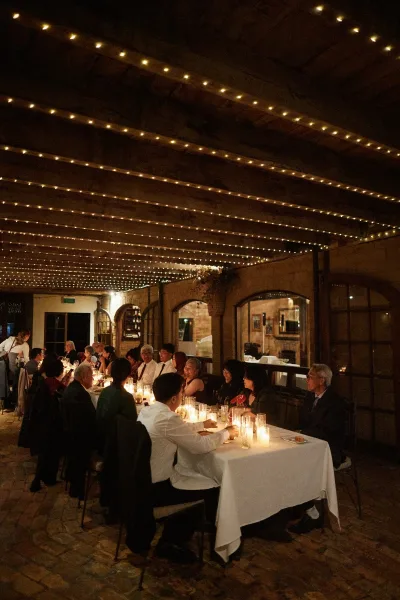 Candlelit reception under string lights in a rustic hall with a wood beam ceiling and arched windows, creating a warm, intimate dinner setting.
