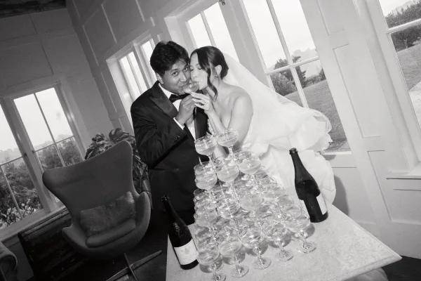 Champagne tower moment with the couple at a wedding champagne tower, the bride’s veil aglow by large windows overlooking a garden lawn.