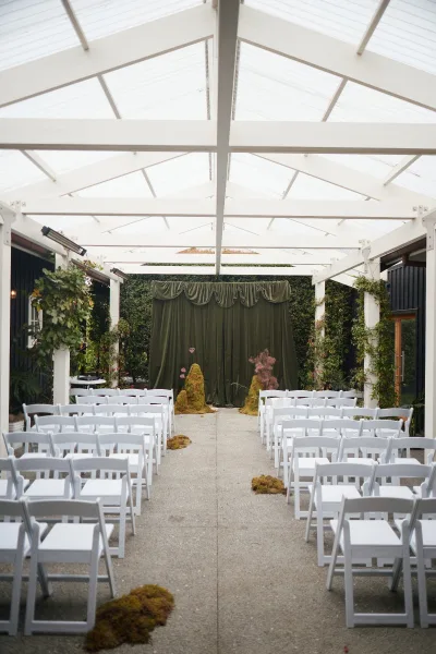 Ceremony setup with moss aisle decor under a pergola’s translucent ceiling and white beams, framed by a neat hedge wall backdrop.
