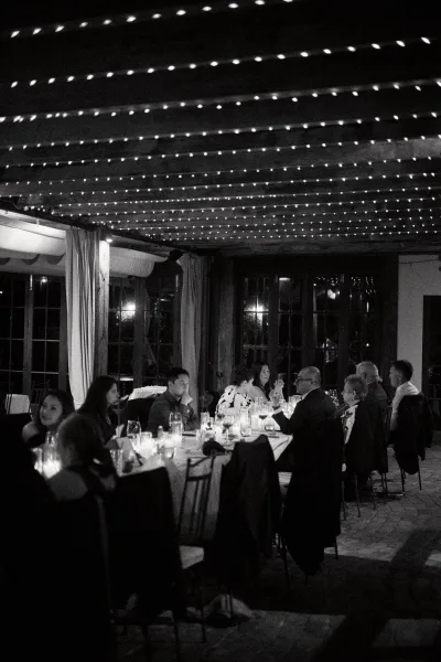 Reception moment at a wedding reception dinner, guests chatting under string lights beneath wood beams and beside large windows.