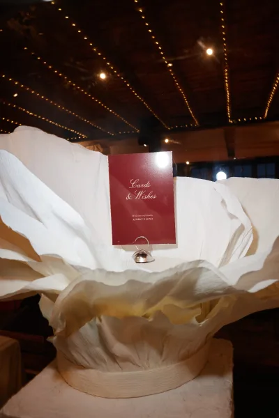 Wedding cards sign inviting cards and wishes, trimmed with a large paper flower, set in an indoor venue beneath string lights and a wooden ceiling.