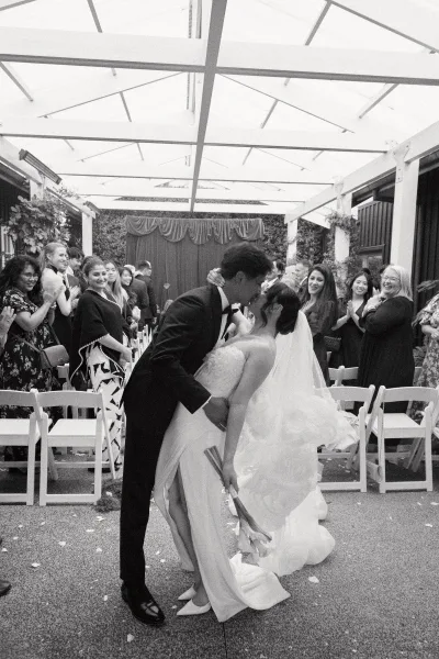 Wedding kiss portrait during a ceremony recessional moment as the groom dips the bride in a covered courtyard aisle, her bouquet of long stems.