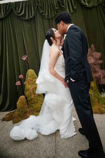 Wedding kiss portrait as a bride and groom embrace, her ruffled train sweeping the concrete floor before a velvet drape.