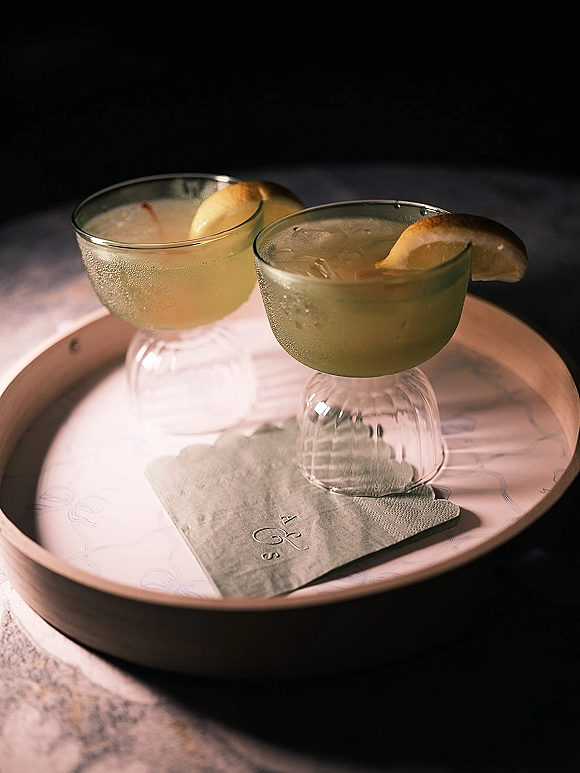 Wedding cocktails in coupe glasses with lemon wedges and ice on a serving tray, set on a dark tabletop with moody shadows