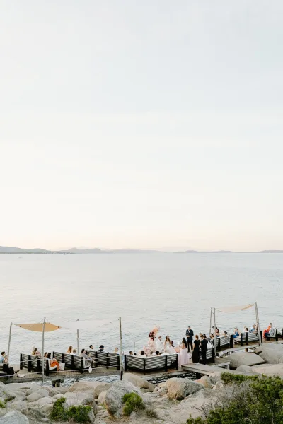 Ceremony moment at a waterfront wedding ceremony beneath shade sails, with water and rocky shoreline to distant hills