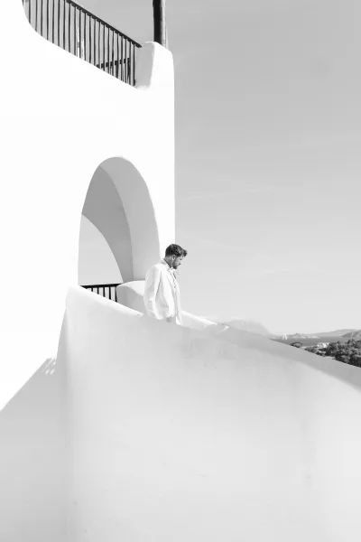 Groom portrait in a sleek suit on a white stucco staircase beneath an archway, with balcony railing, sky, and distant hills.