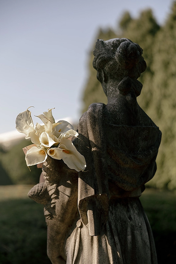 Calla lily bouquet of white blooms with ribbon wrap resting on a stone statue in an outdoor garden with trees and sky