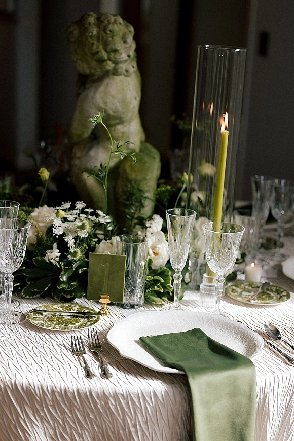 Reception tablescape with green wedding tablescape accents—taper candle, crystal glasses, green napkin, white florals and garland in a dim room
