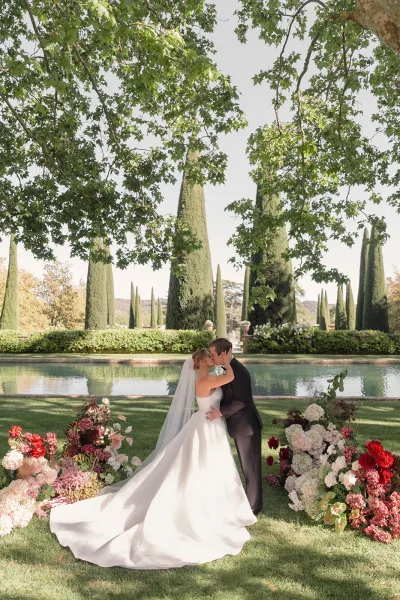 Wedding kiss portrait as the bride and groom kiss by a reflecting pool, framed by cypress trees, hedges, and colorful floral arrangements.