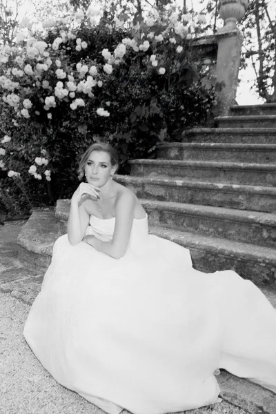 Bridal portrait of a bride in a strapless wedding dress, seated on stone steps amid rose bushes, with delicate drop earrings enhancing the look.