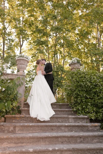 Wedding kiss portrait on a stone staircase, a staircase kiss as the couple embraces amid sunlit garden greenery and trailing vines.