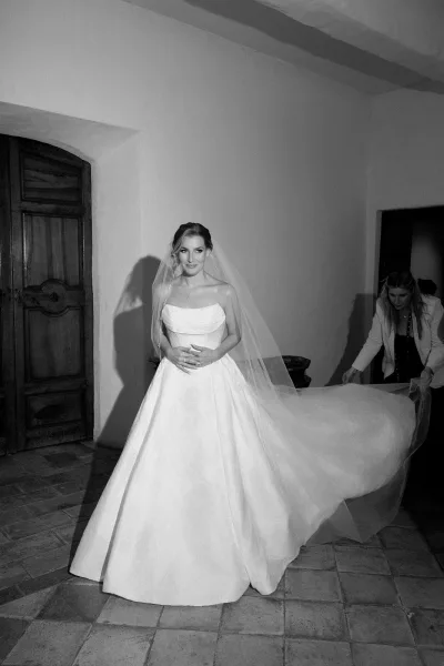 Bridal portrait of a bride getting ready, her veil draping as she pauses by a wooden door in a quiet indoor hallway.