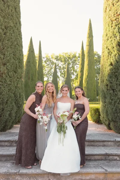 Bride and bridesmaids pose for a garden bridal portrait on stone steps flanked by cypress trees, each carrying pastel bouquets.