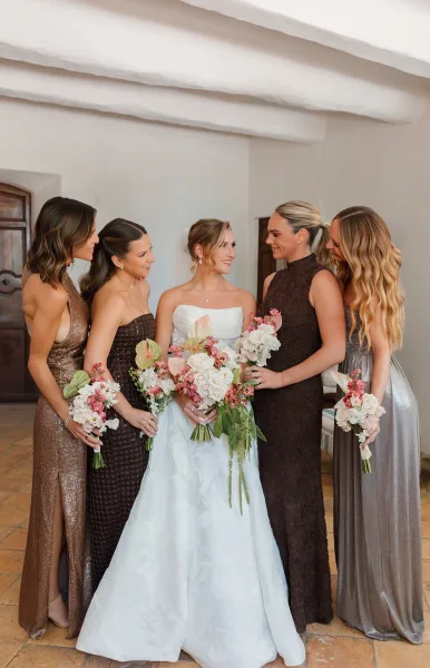 Bride and bridesmaids holding white and blush bouquets stand against white plaster walls and rustic wooden doors.