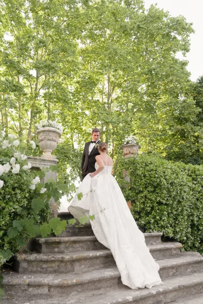 Couple portrait of a bride and groom on stairs, her ballgown train sweeping a garden stone staircase accented by white florals and greenery.