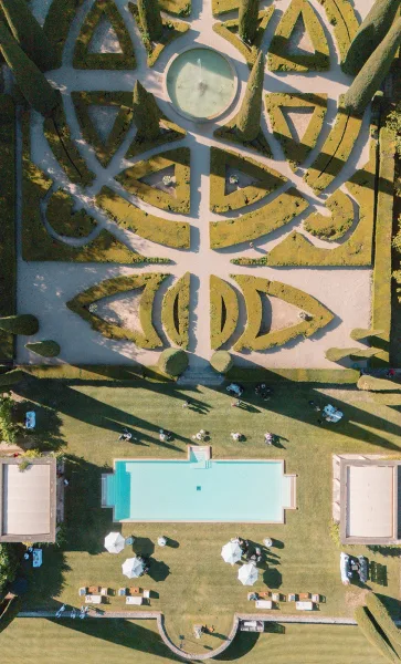 Poolside reception setup arranged for an outdoor cocktail hour, with white umbrellas shading lounge areas beside a formal garden lawn.