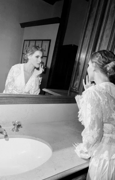 Bride getting ready during a bridal makeup moment, applying lipstick before a mirror at a wood-paneled bathroom vanity.