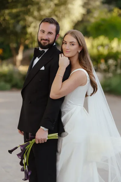 Couple portrait of bride holding groom’s arm as he holds a calla lily bouquet, her long veil flowing in a lush garden walkway