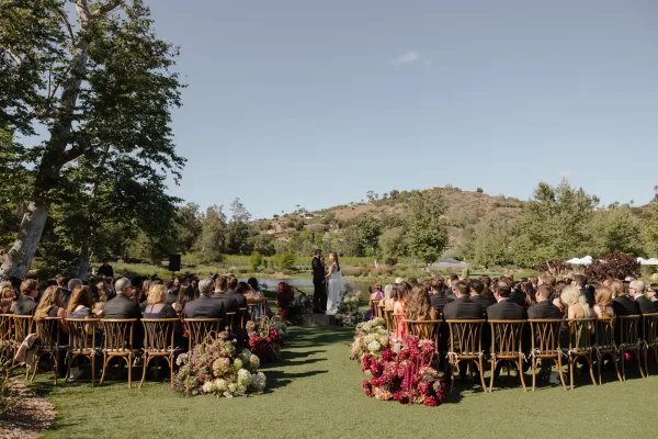 Ceremony moment with aisle florals at an outdoor wedding ceremony, viewed from the back, with wooden chairs and a lake and hills beyond