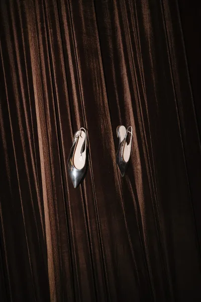 Bridal shoes in a wedding shoe flatlay, silver slingback pointed-toe heels resting on a dark brown velvet drape with rich texture