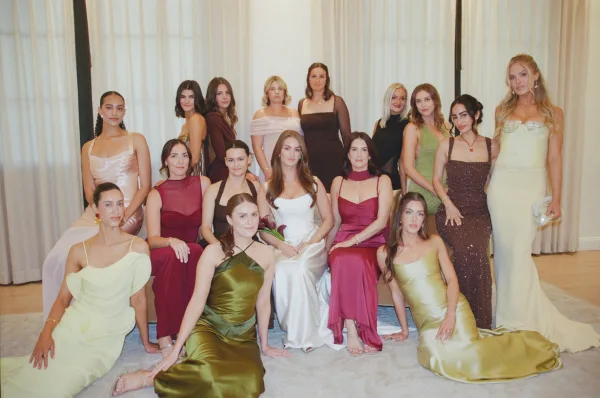 Bridesmaid group photo with bride in strapless gown, bridesmaids in mixed satin and sequined dresses posing by white curtains indoors