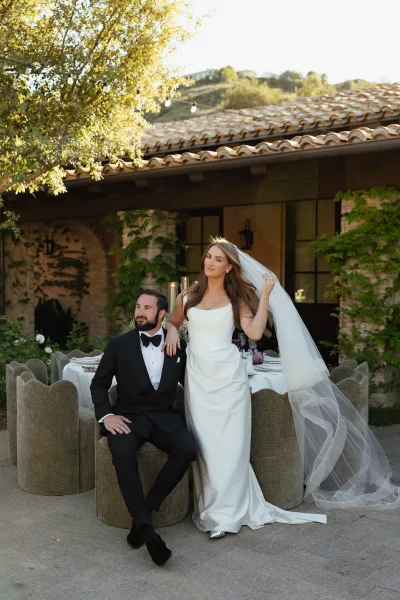 Couple portrait of bride in simple satin wedding dress with cathedral veil beside groom in black tuxedo on courtyard patio under string lights