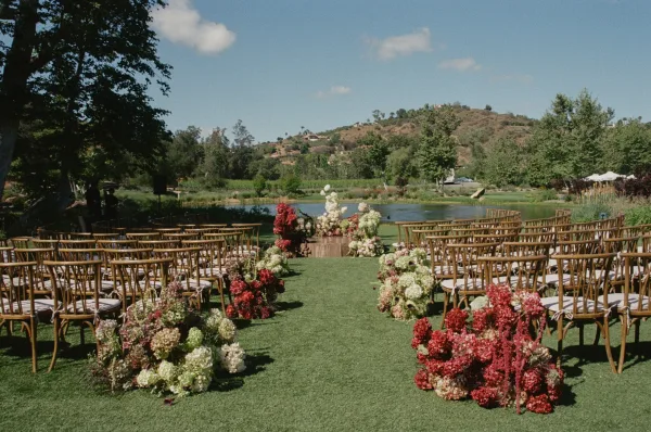 Ceremony aisle decor with hydrangea floral clusters lining a green turf runner, wood chairs facing a lakeside backdrop of trees and hills