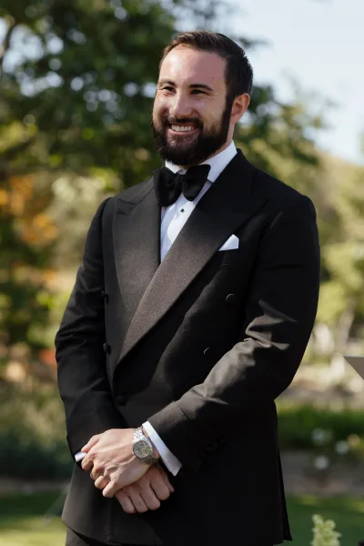 Groom portrait in a black tuxedo with bow tie, pocket square, and wristwatch, smiling outdoors among trees and greenery in daylight
