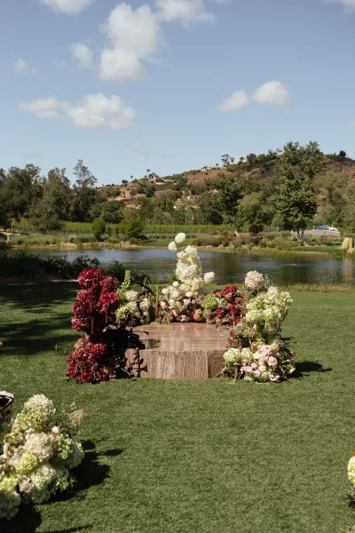 Ceremony altar decor with hydrangeas and blush-burgundy florals on a platform, set on a lawn by a pond with hills and blue sky