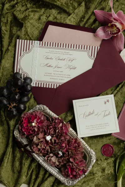 Wedding invitation suite with burgundy wedding invitations, lace ribbon and wax seal, orchids and rings arranged on green velvet fabric