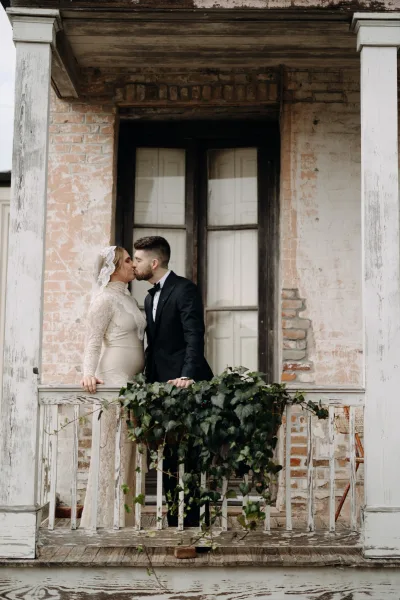 Wedding kiss as bride in long sleeve lace dress and veil leans into tuxedo groom by ivy garland railing on a weathered porch