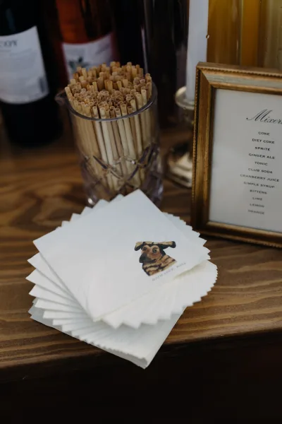 Wedding bar signage in a gold frame with signature cocktail menu sign, paper napkins, dog sticker, straws, and liquor bottles on wood tabletop