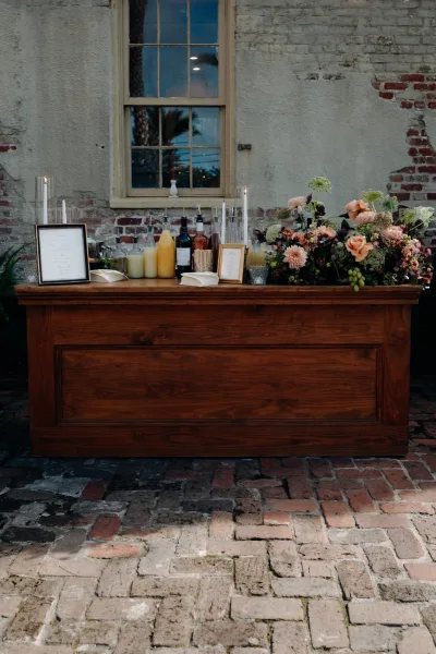 Wedding bar setup with liquor bottles, glassware, and taper candles on a wood bar, framed menu and florals against stucco and brick wall