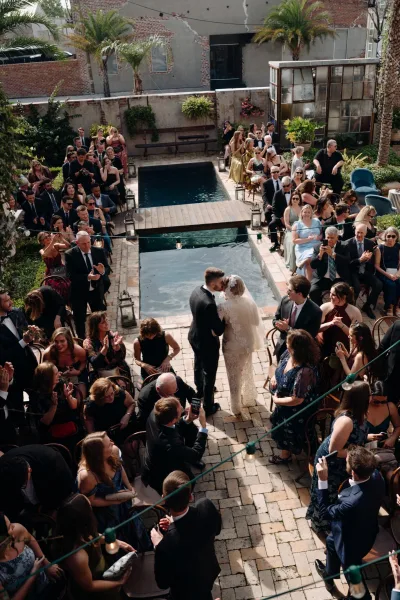 Ceremony kiss as bride in a lace dress and veil and groom in tuxedo embrace beside a pool in a brick courtyard under string lights