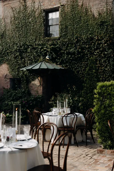 Outdoor reception tables set with white linens, bentwood chairs, and glass cylinder taper candles in an ivy-covered brick courtyard
