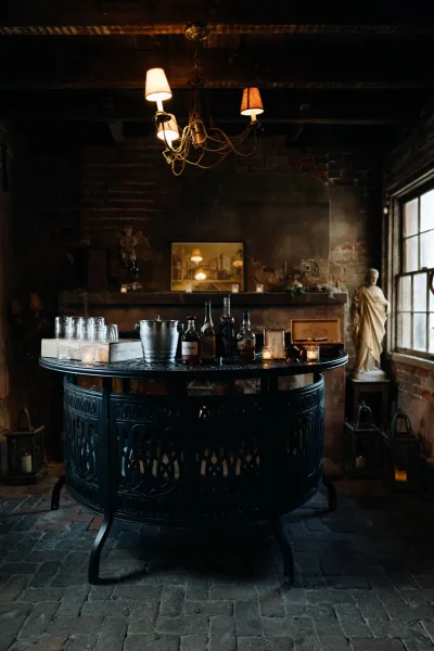 Wedding bar setup with round bar table, liquor and wine bottles, glassware, and candles against a rustic brick wall under a chandelier