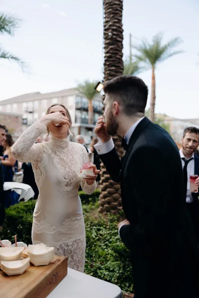 Wedding reception moment as bride in a high neck lace gown and groom in tuxedo share dessert beside cocktail glasses in a palm courtyard