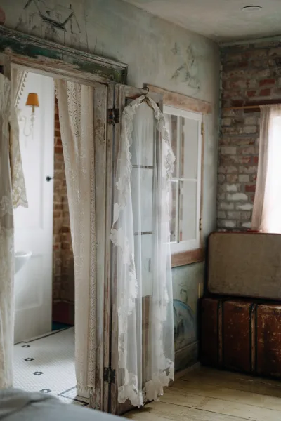 Bridal veil with lace wedding veil trim hangs from a wooden hanger by a brick wall window, sheer tulle draped over a vintage suitcase